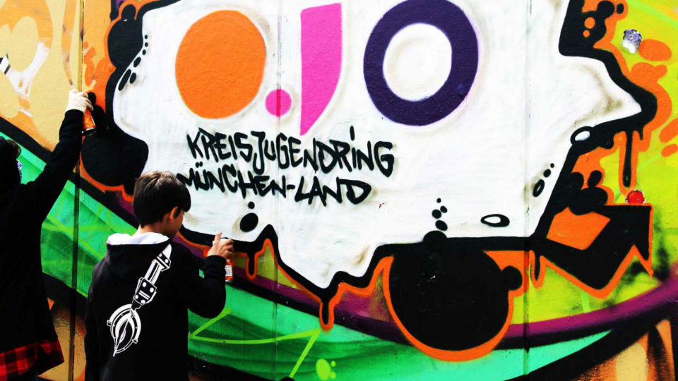 Zwei Kinder sprayen auf eine Wand ein Graffiti mit dem Schriftzug OJO Kreisjugendring München-Land