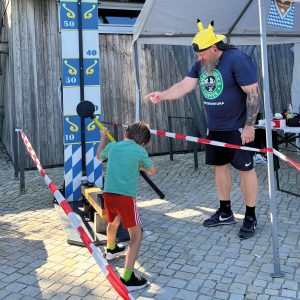 Ein Junge haut den Hammer auf das Jahrmarktsgerät "Hau den Lukas". Ein Mitarbeiter hilft ihm und gibt Tipps. 