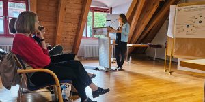 Frau steht am Renderpult des Landratsamts, hinter ihr stehen Pinnwände mit Plakaten. Das Publikum hört zu.