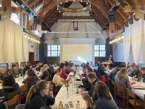 Ansicht der Delegierten an drei großen Tischreihen, Vorsitzender Daniel Gögelein spricht am Rednerpult und auf der Leinwand ist zu lesen "Politische Bildung - Kurzvortrag"