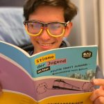Ein Teilnehmer liest mit einer Deutschlandbrille das Reisetagebuch zur Fahrt und lacht.